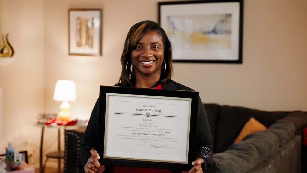 A smiling woman holds a framed nursing license. She stands in a warmly lit living room with framed artwork and a lamp in the background.