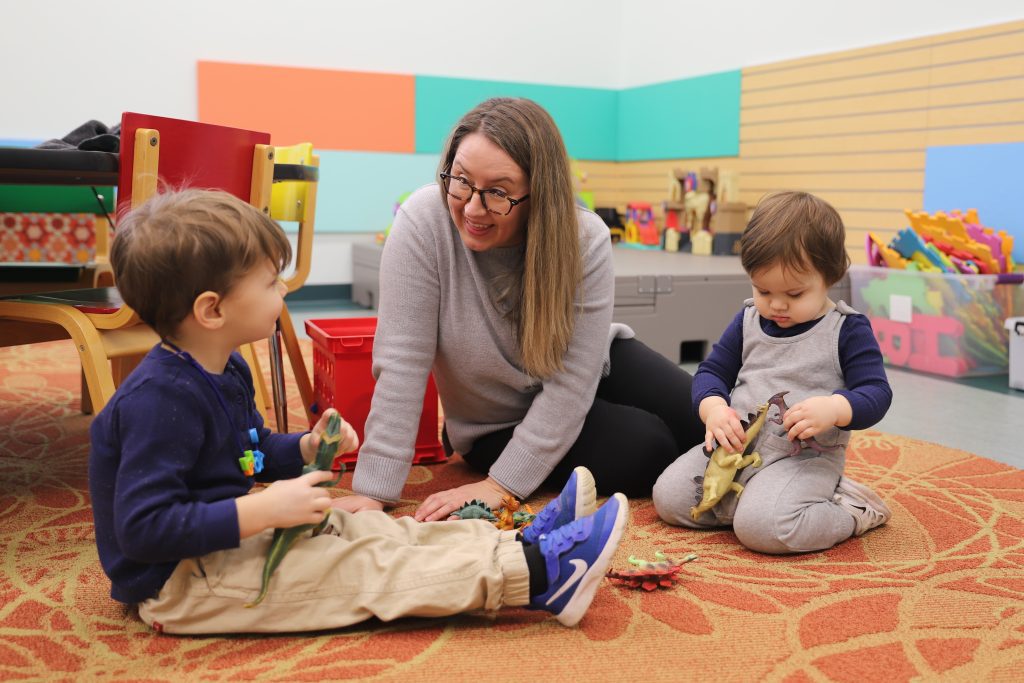 A mother with glasses and long blonde hair kneels on the floor, smiling and engaging with two young children playing with toy dinosaurs in a colorful classroom. The children, a boy in a navy blue shirt and beige pants and a toddler in gray overalls, sit on a patterned rug surrounded by toys.