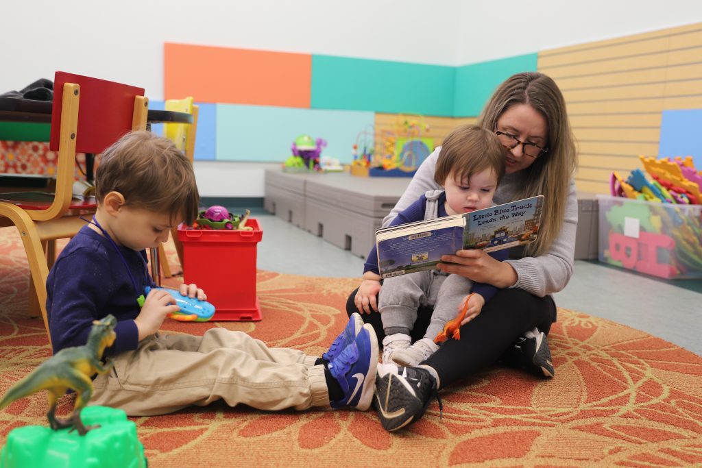 The mother sits on the floor holding a young child in her lap while reading a book titled Little Blue Truck Leads the Way. The child looks intently at the book while another child in the foreground plays with a toy in the same classroom setting.