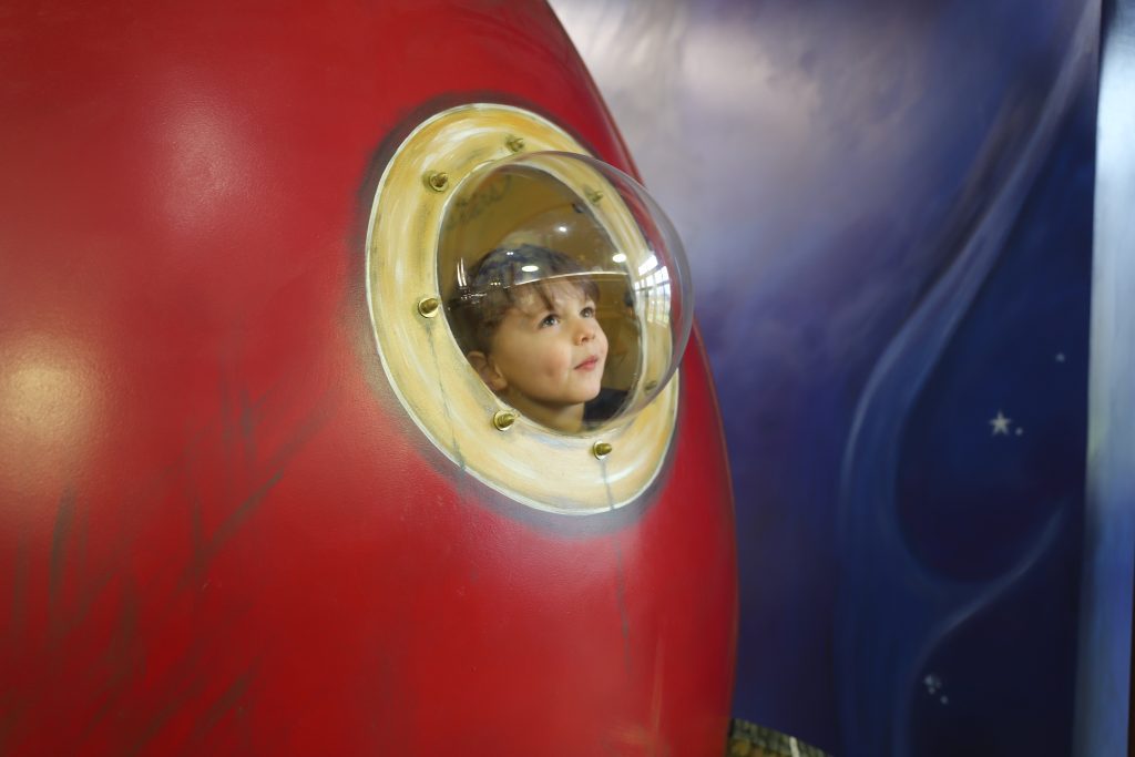 A young boy gazes out of a circular window in a red play structure resembling a spaceship. The background is painted with a cosmic theme, featuring stars and swirling blue hues.