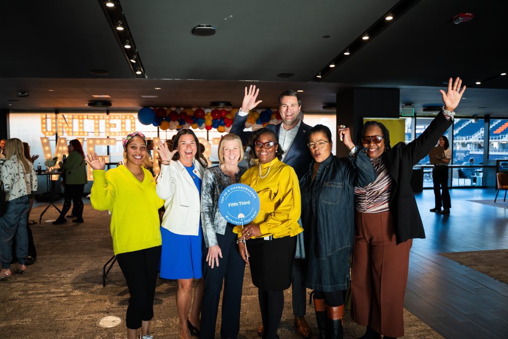 A diverse group of seven people is posing enthusiastically at an indoor event, smiling and raising their hands. One person in the center holds a circular sign that reads "Fifth Third" with additional text. The background features a lit-up sign spelling "UNITED WE THRIVE" along with colorful balloons and a modern venue with large windows overlooking a stadium.