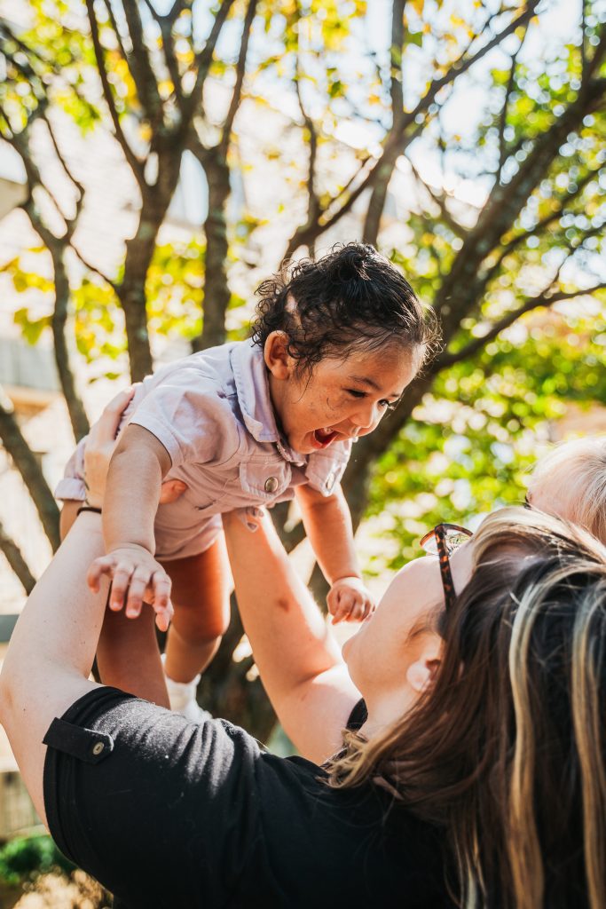 A woman joyfully lifts a smiling toddler into the air on a sunny day, with trees and sunlight creating a warm, playful backdrop. The child looks excited and happy, reaching forward with laughter.
