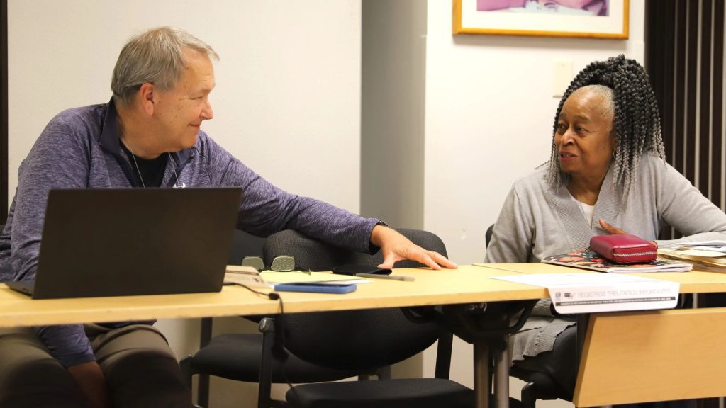 Two individuals are seated at a table in a casual meeting setting, engaged in a friendly conversation. The man on the left has a laptop open in front of him, while the woman on the right has magazines, a folder, and a red pencil case on the table. Both are smiling, creating a warm and collegial atmosphere.
