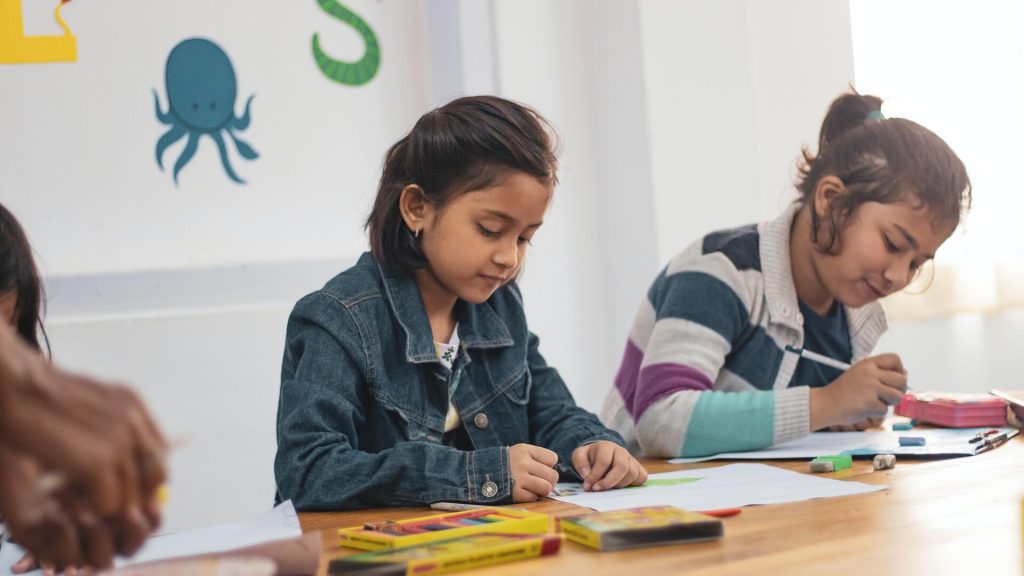 Two young girls are seated at a table engaged in a creative activity, drawing or coloring with crayons and pencils. The classroom setting is bright and cheerful, with playful wall decorations including an octopus. The children appear focused and content as they work on their artwork.