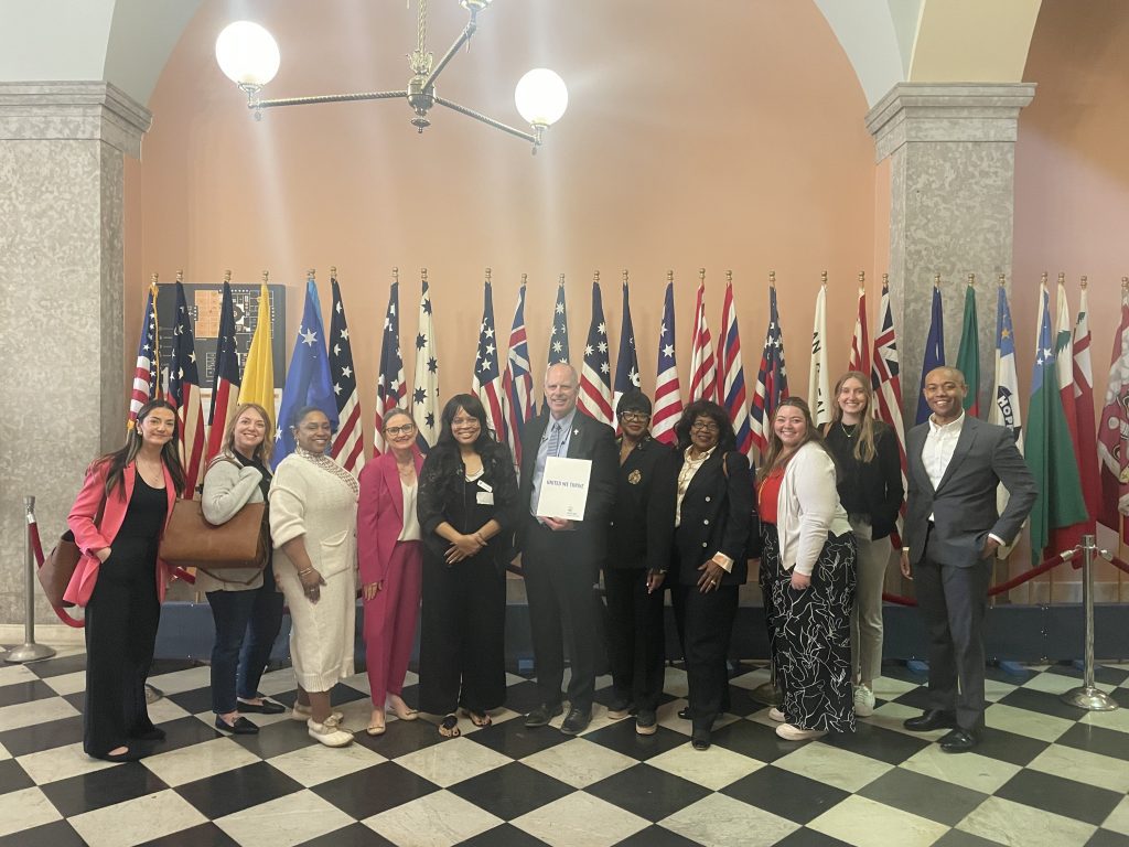 A diverse group of thirteen people is gathered in the Ohio Statehouse with checkered flooring and a backdrop of numerous international and U.S. flags. They are posing and smiling for the camera, with one person in the center holding a folder labeled "UNITED WAY WE THRIVE." The group stands beneath a grand chandelier in a building that appears to be a government or civic space.