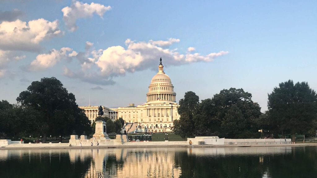 The U.S. Capitol building in Washington, D.C., is shown at sunset with a few scattered clouds in the sky. The dome is prominently illuminated by the golden hour light, and its reflection can be seen in the water of the Capitol Reflecting Pool in the foreground. Trees and statues frame the scene, capturing a peaceful and iconic view of the nation's capital.