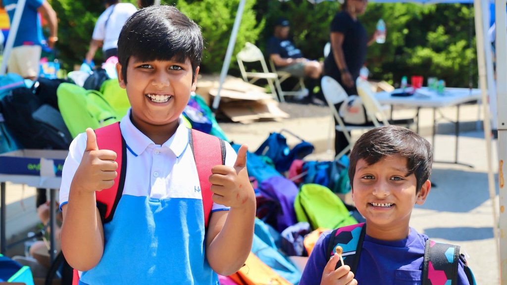 Two young boys wearing backpacks smile brightly at the camera, one giving a double thumbs-up and the other holding a lollipop, with colorful school supplies and volunteers visible in the background at an outdoor distribution event.