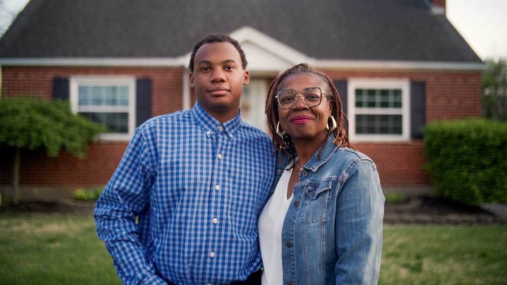 A mother and son stand smiling in front of their home.