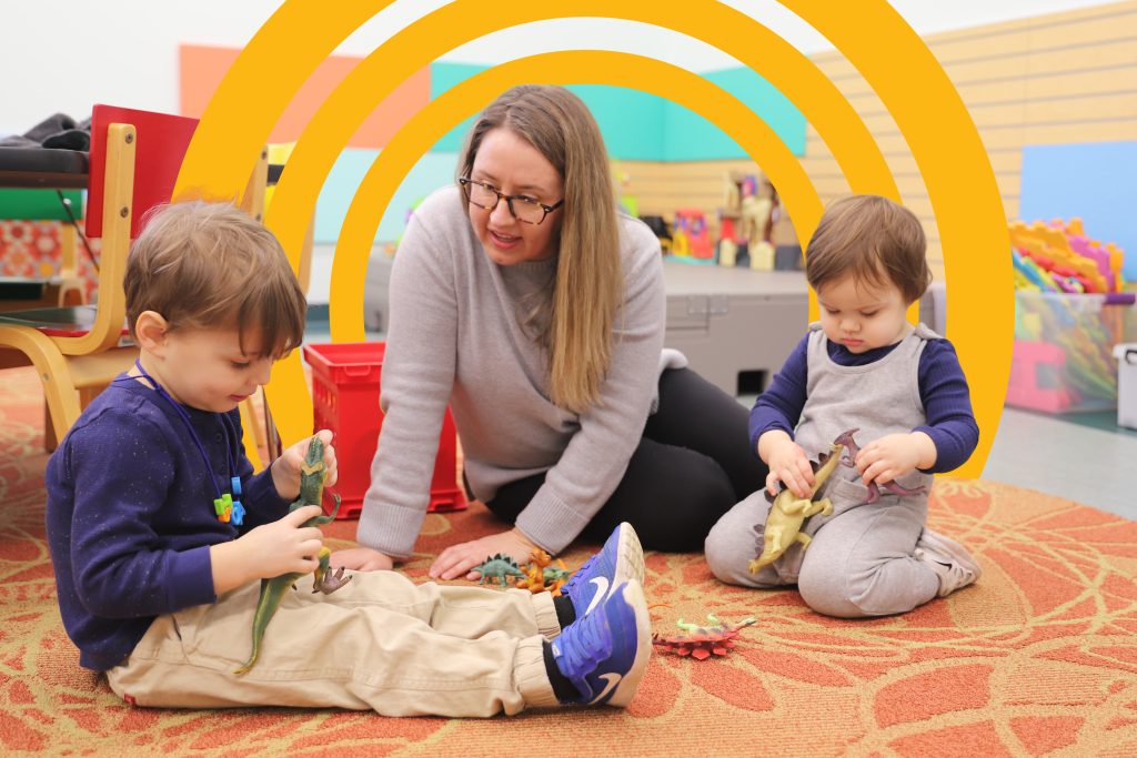 A mother with glasses and long blonde hair kneels on the floor, smiling and engaging with two young children playing with toy dinosaurs in a colorful classroom. The children, a boy in a navy blue shirt and beige pants and a toddler in gray overalls, sit on a patterned rug surrounded by toys.