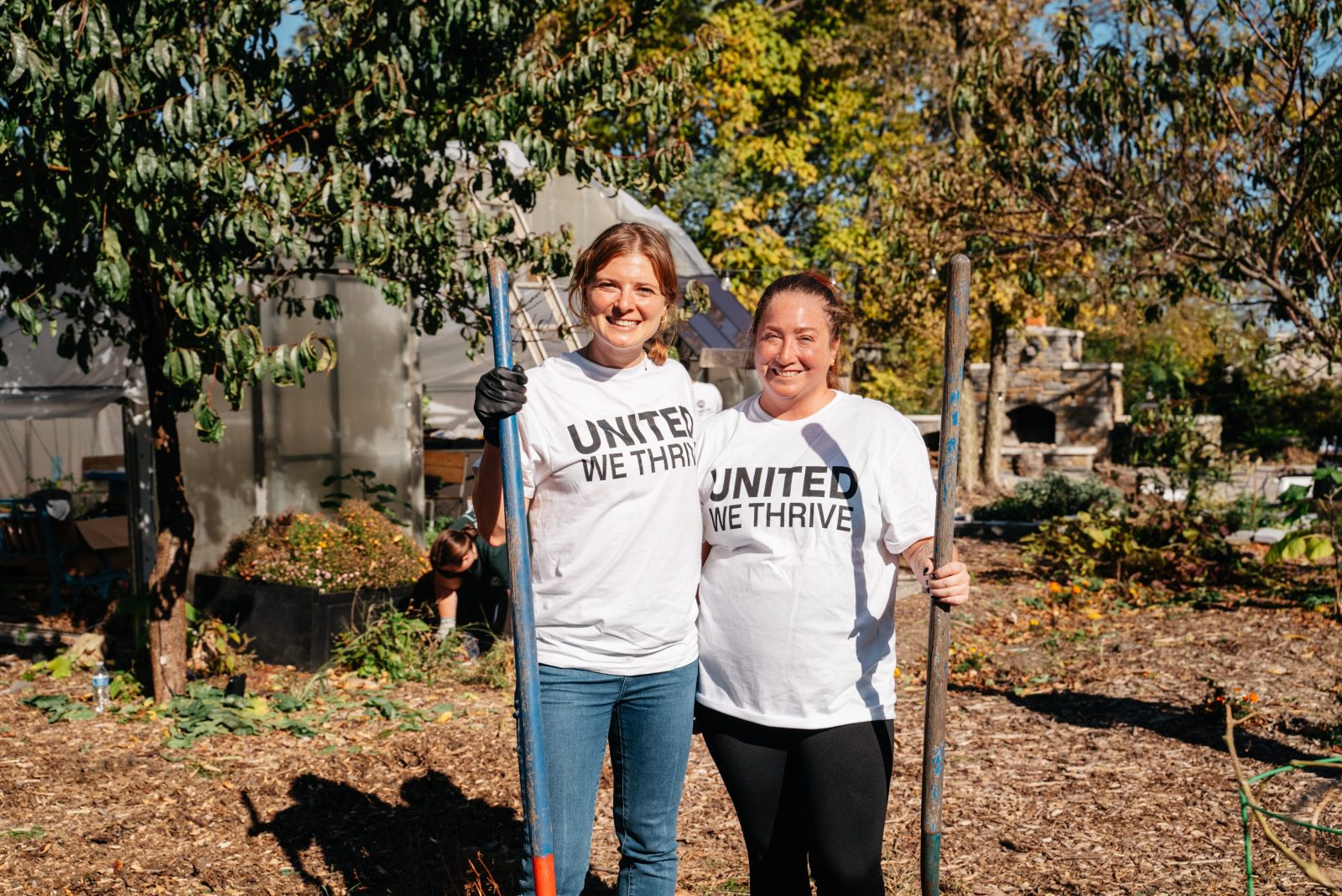 Two people stand side by side in a garden holding tools, wearing white T-shirts that say "United We Thrive.