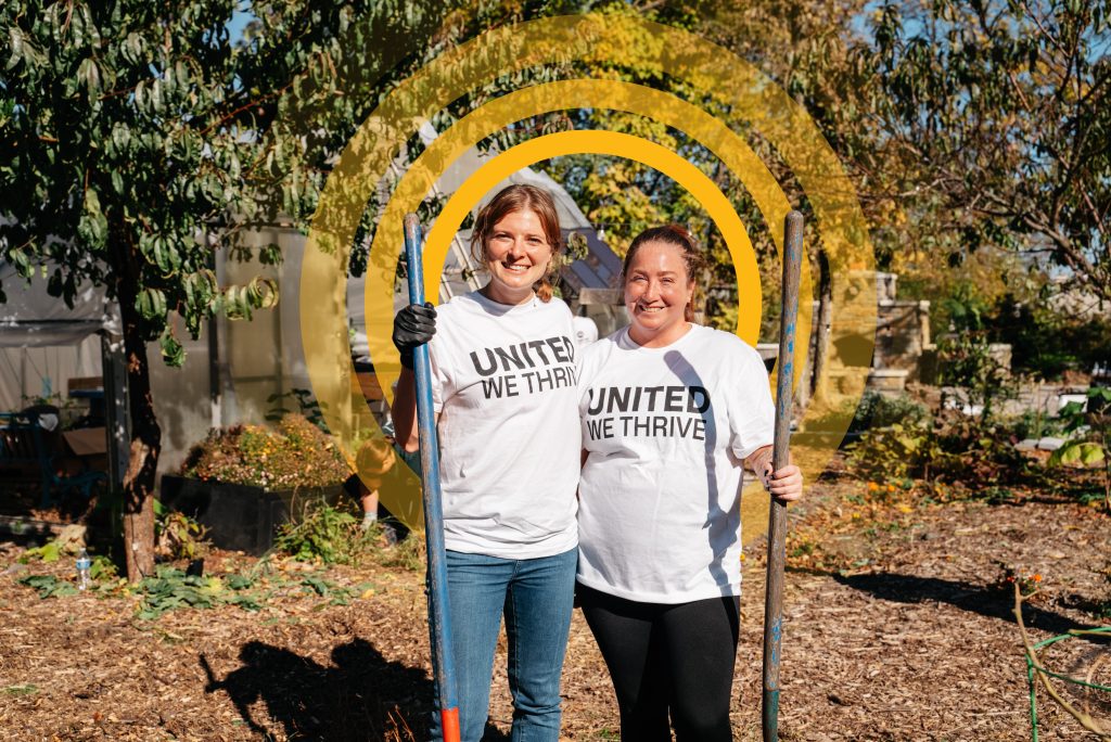Two people stand side by side in a garden holding tools, wearing white T-shirts that say "United We Thrive.