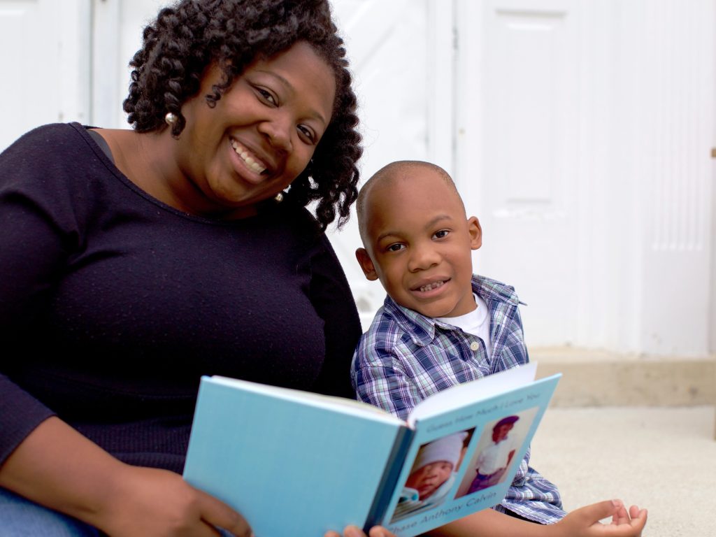 Adult and young child sitting on steps, smiling at the camera while holding an open book together.