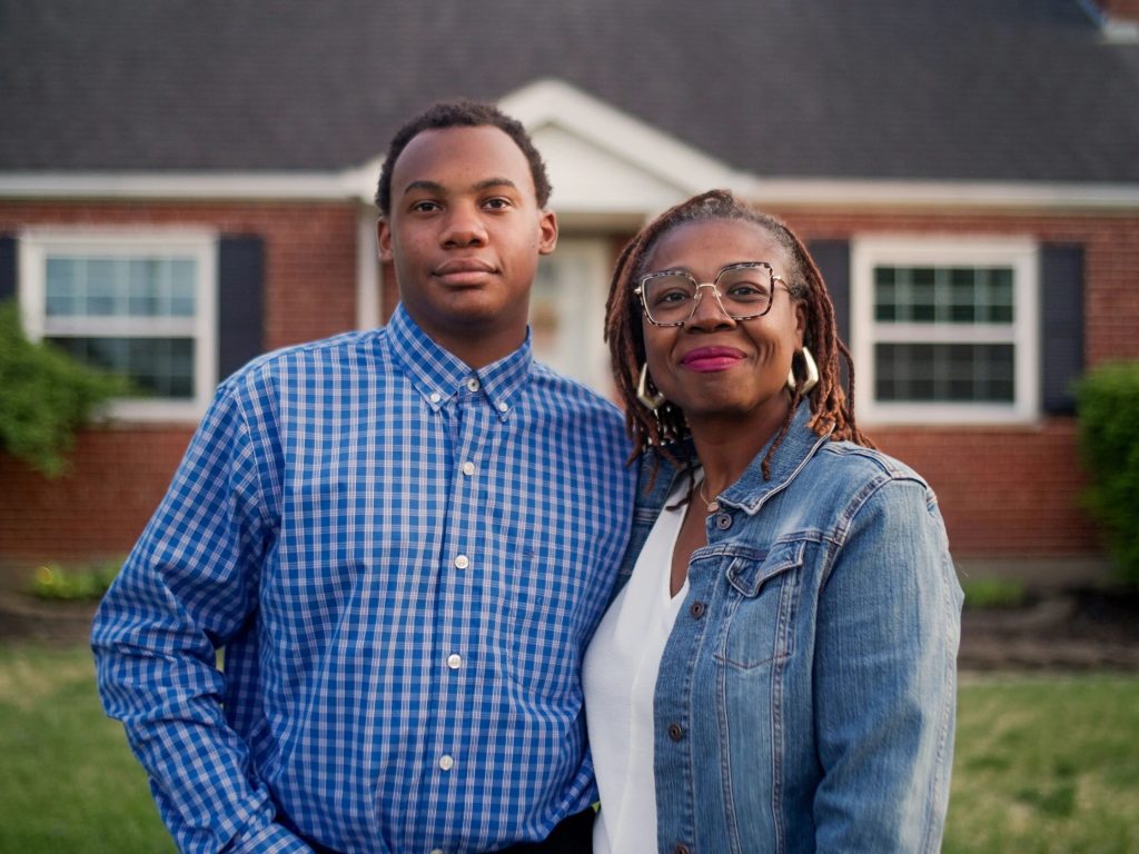 Two people standing side by side outdoors in front of a brick house, looking at the camera.