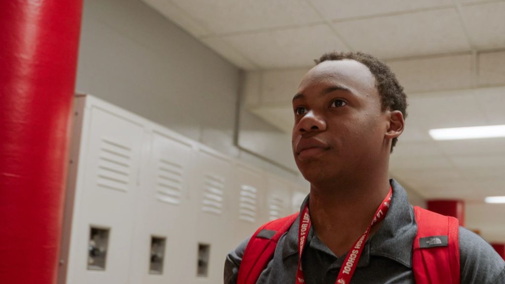 A person stands in a hallway with lockers, wearing a red lanyard and carrying a red backpack.