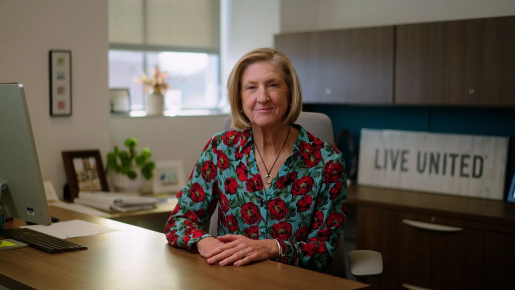 A person sits at a desk in an office, wearing a floral shirt. The desk has a computer monitor and papers, with a 'LIVE UNITED' sign visible on a shelf behind.