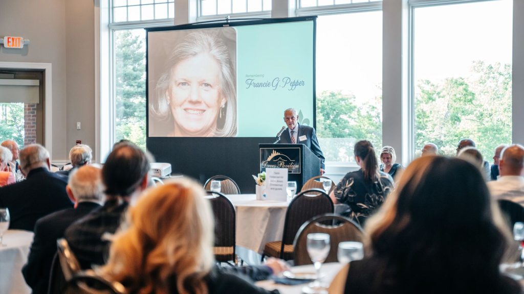 A speaker stands at a podium in front of a seated audience during an event, with a large screen behind him displaying a photo and the name "Francie G. Pepper."