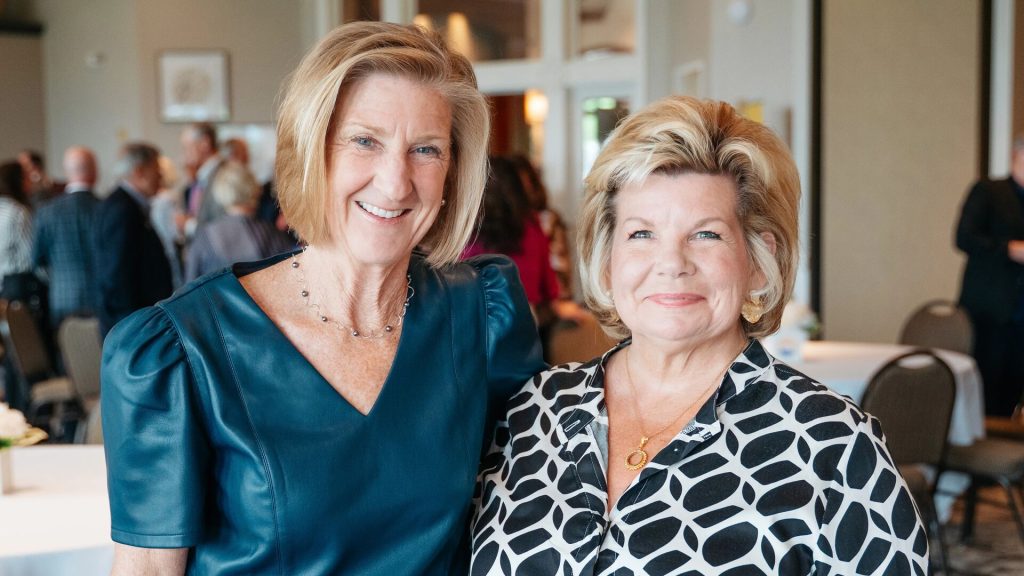 Two people stand close together in a room set up for an event, with tables, chairs, and framed pictures on the walls. One wears a blue dress, the other a black and white patterned blouse.