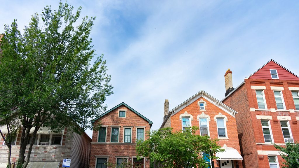 A row of houses with varied architectural styles, a tree on the left, and a clear sky with light clouds.