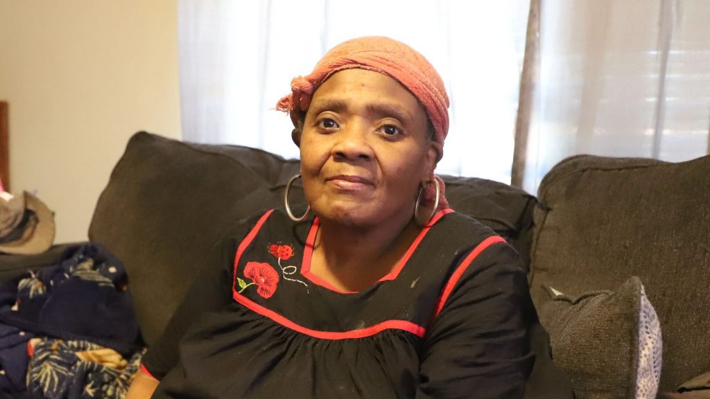 Person wearing a black top with red trim and floral embroidery, and a pink headscarf, sitting on a dark-colored couch. Light-colored curtains with sunlight in the background; clothing items are visible on the left side of the couch.