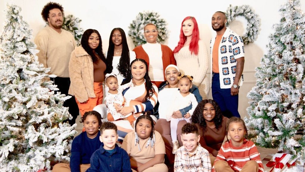 Group of adults and children posing in front of a festive holiday backdrop with decorated Christmas trees and wreaths.