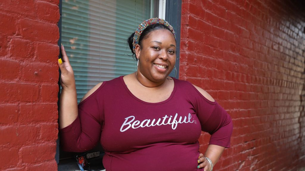 Person in a maroon shirt with 'Beautiful' written in white cursive, standing against a red brick wall with one hand on the wall and the other on their hip.