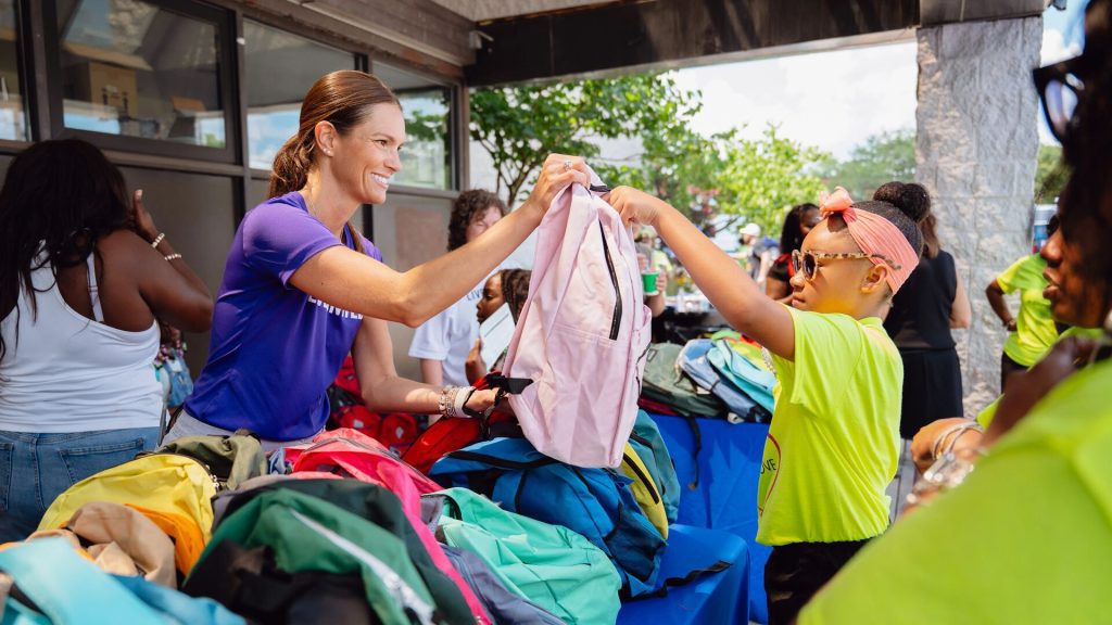 Volunteers distribute colorful backpacks at an outdoor community event, with one person in a purple shirt handing a pink backpack to a child in a yellow shirt, surrounded by tables and trees.