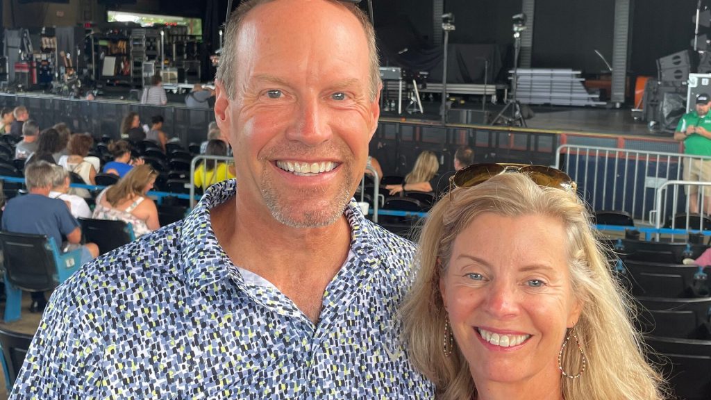 Two people standing in front of a stage at a concert or event venue, with attendees seated in the background and musical equipment visible on stage.