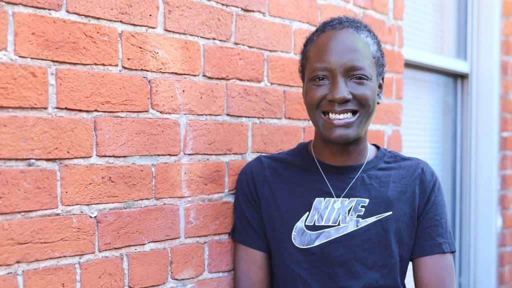 "Person standing in an alleyway against a brick wall, wearing a black Nike t-shirt with a visible logo.