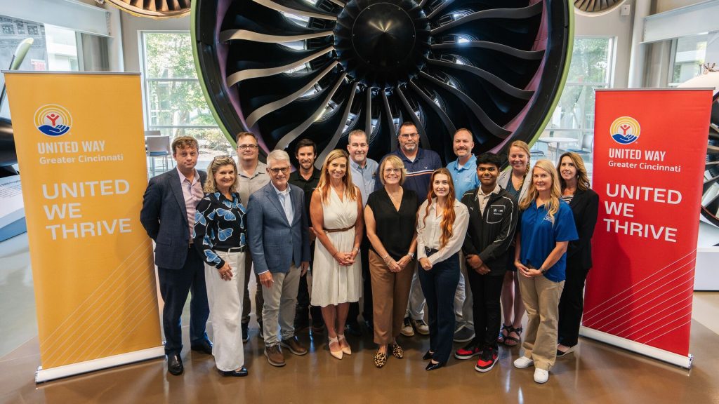 Group of people in business casual attire posing in front of a large jet engine display, flanked by banners reading 'UNITED WAY Greater Cincinnati' and 'UNITED WE THRIVE.'