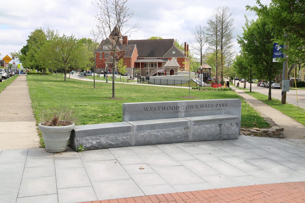 Stone bench engraved with 'WESTWOOD TOWN HALL PARK' in a grassy park area, with a red brick building and trees in the background under a partly cloudy sky.