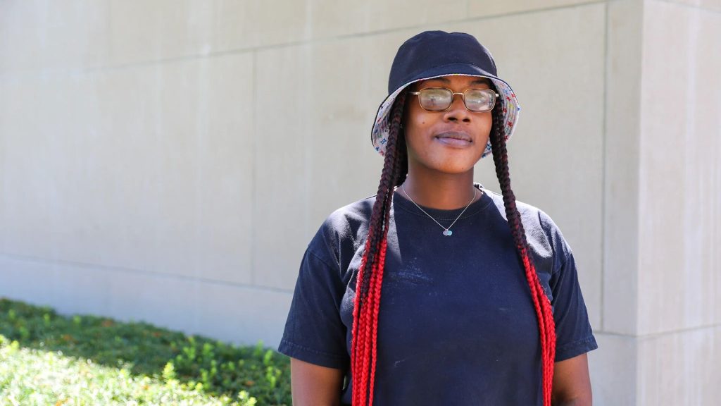 Person wearing glasses and a bucket hat, standing outside in front of a light-colored wall with greenery in the foreground.