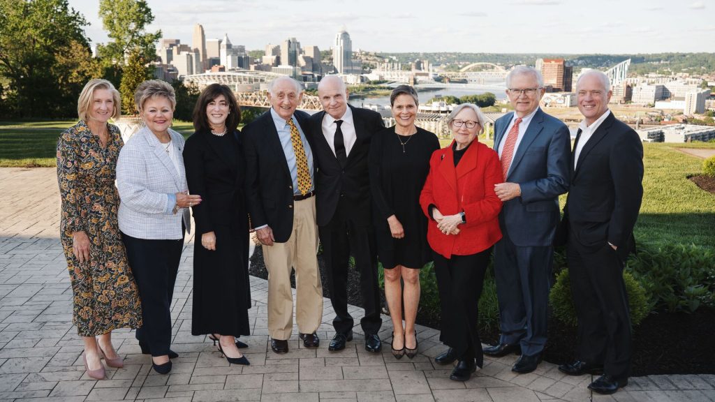 Nine adults in formal attire stand together outdoors with a city skyline and river in the background.