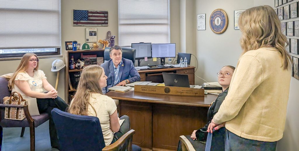 Group meeting in a legislative office with three young women and one woman speaking to a seated representative behind a desk.