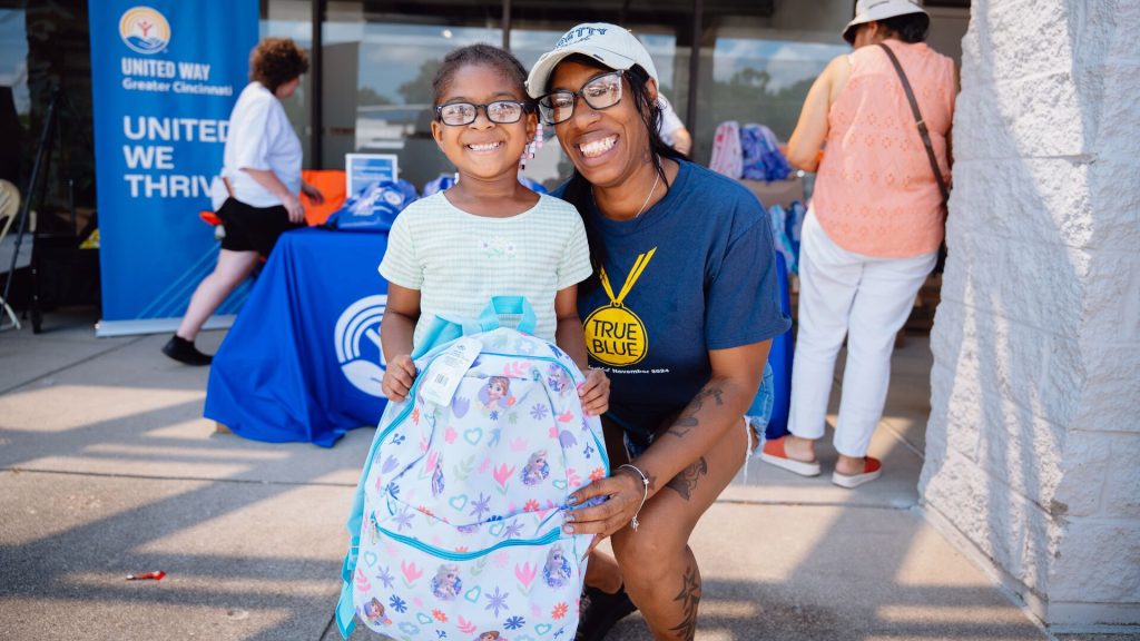 A young girl in glasses smiles holding a Frozen-themed backpack with a woman kneeling beside her at a United Way of Greater Cincinnati backpack giveaway event