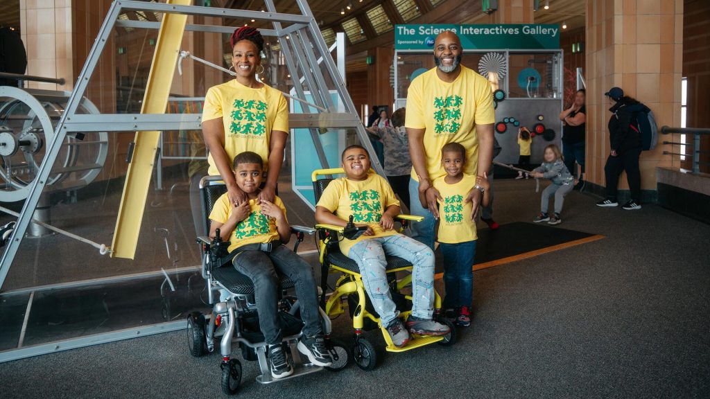 A family wearing yellow shirts walks through a large train station hall, with a child in a wheelchair labeled “MATRIX Ultra” and a mural on the wall in the background.