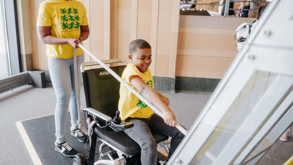 A child in a wheelchair labeled “MATRIX” smiles while holding a rope, as an adult stands behind in a hallway with tiled walls.