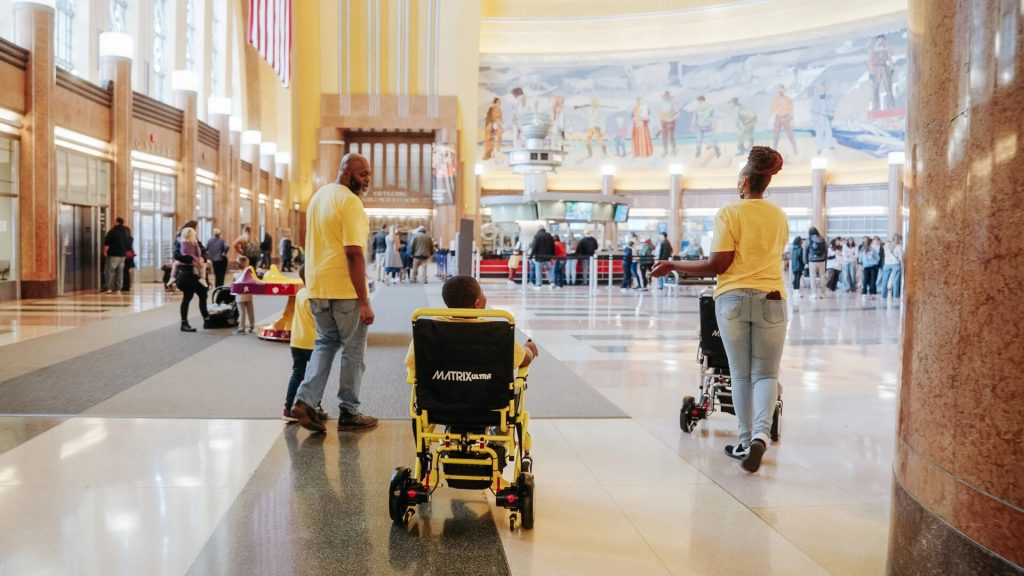 A family wearing yellow shirts walks through a large train station hall, with a child in a wheelchair labeled “MATRIX Ultra” and a mural on the wall in the background.
