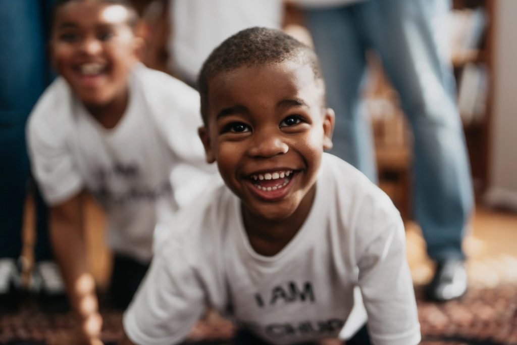 Child smiling and leaning forward toward the camera, with another child visible in the background.