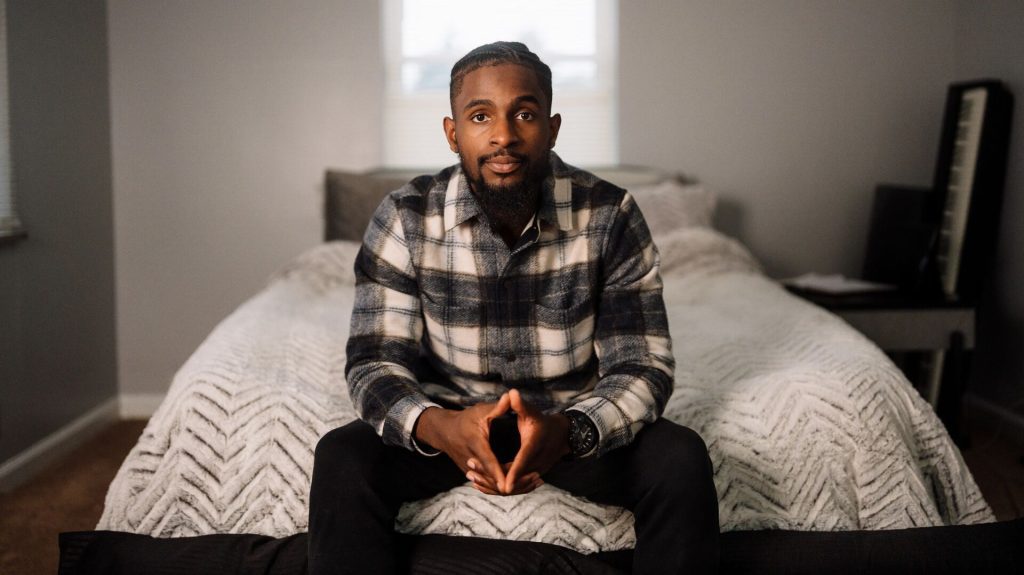 Person sitting on the edge of a neatly made bed, looking at the camera in a softly lit bedroom.