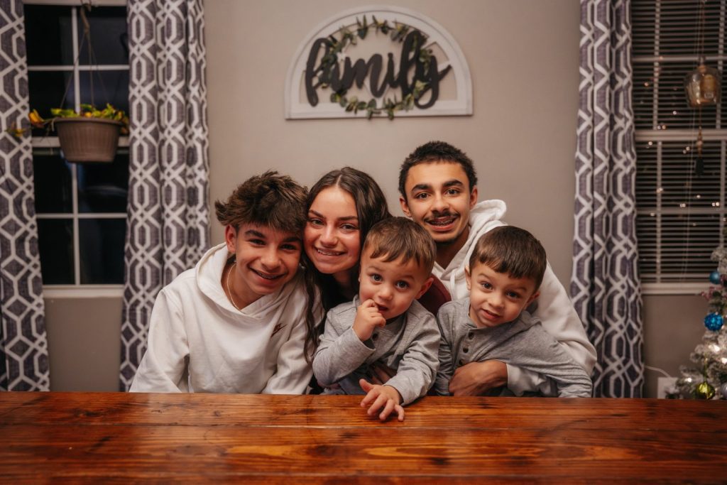 Family sitting together at a wooden table, smiling at the camera in a cozy room with patterned curtains and a “family” sign on the wall.