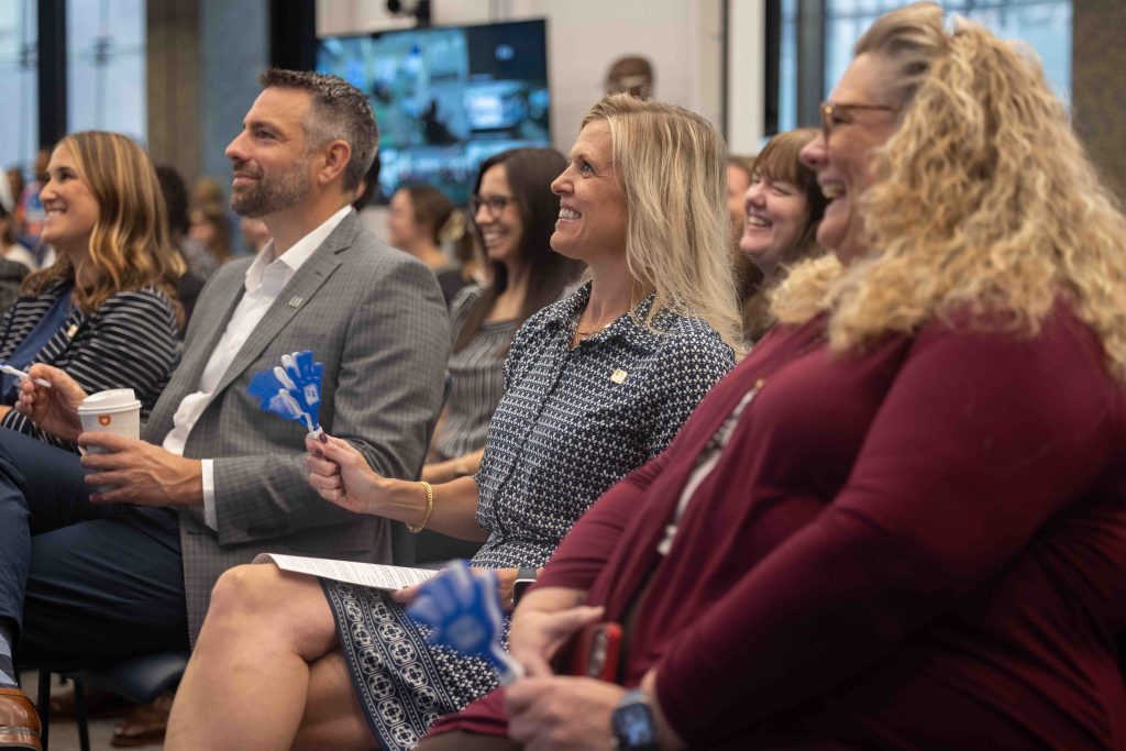 Audience members in business attire sit in rows smiling and holding small blue hand-shaped noisemakers at an indoor event.