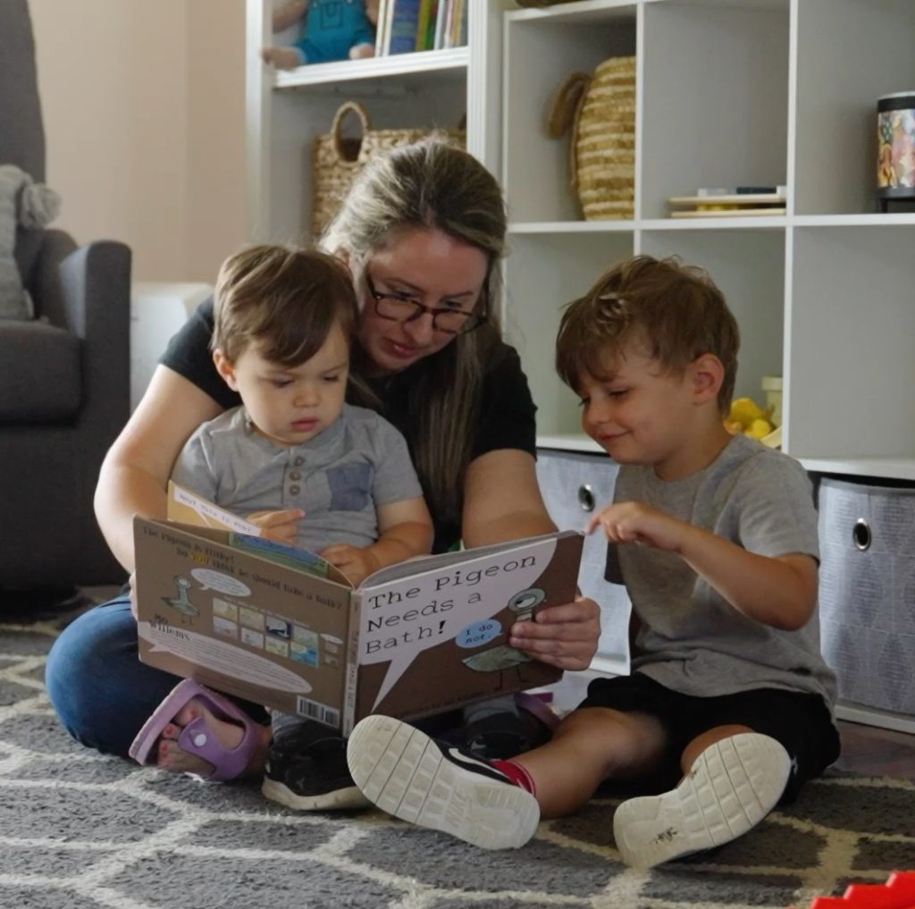 Adult sitting on the floor reading a picture book with two young children in a living room.