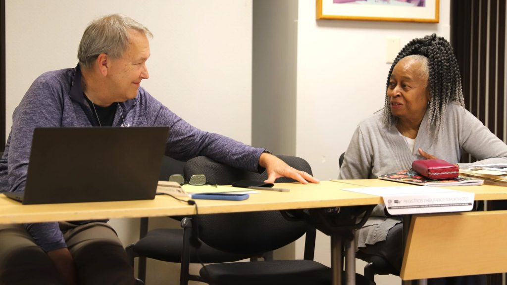 Two people sitting at a table talking, with papers and a laptop in front of them in an indoor setting.