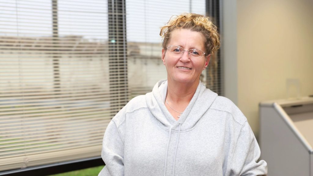 A person with curly hair and glasses wearing a gray hoodie sits in an office cubicle with window blinds in the background.
