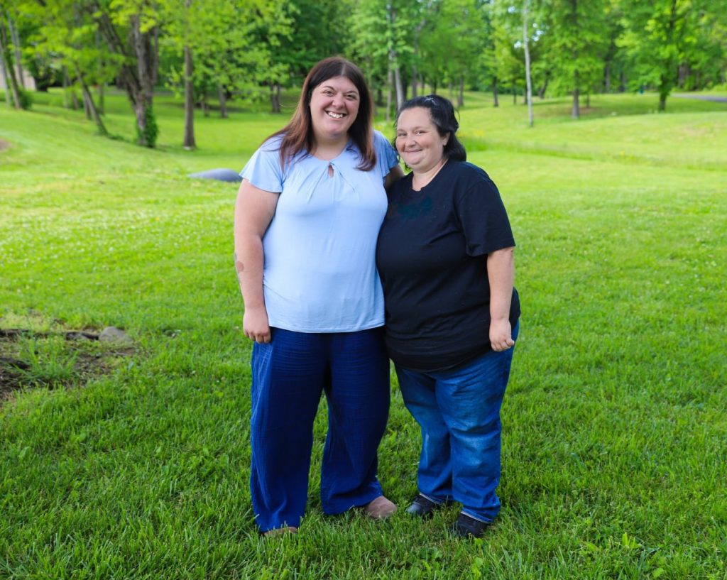 Two women stand together and smile in a grassy park.