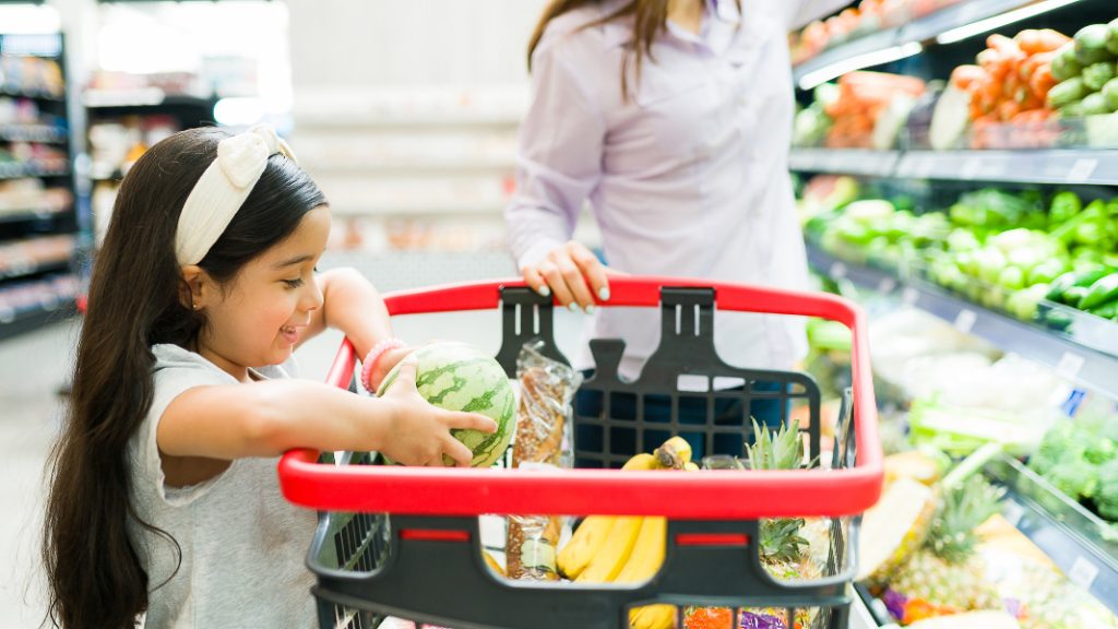 Child smiling while placing a small watermelon into a grocery cart filled with fruits and vegetables, next to an adult in a supermarket produce section.