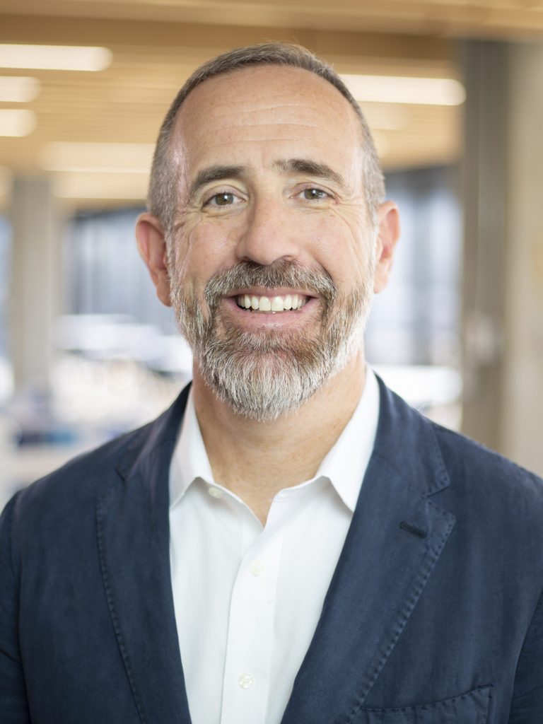 An image of a man in a white collated shirt and blue coat with an office in his background.