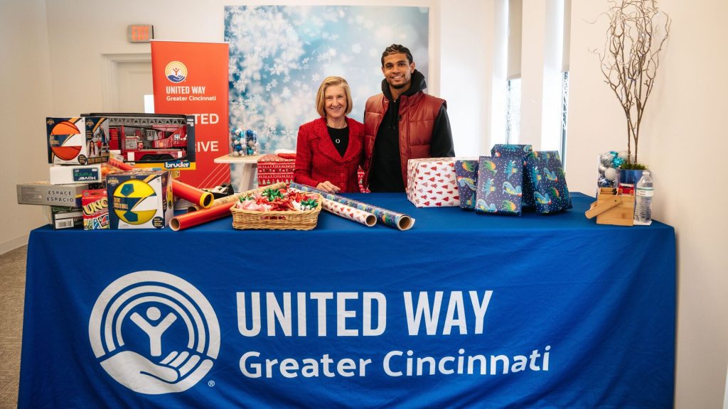 Two people standing behind a table covered with wrapped gifts and toys, with a United Way Greater Cincinnati banner in a decorated indoor space.