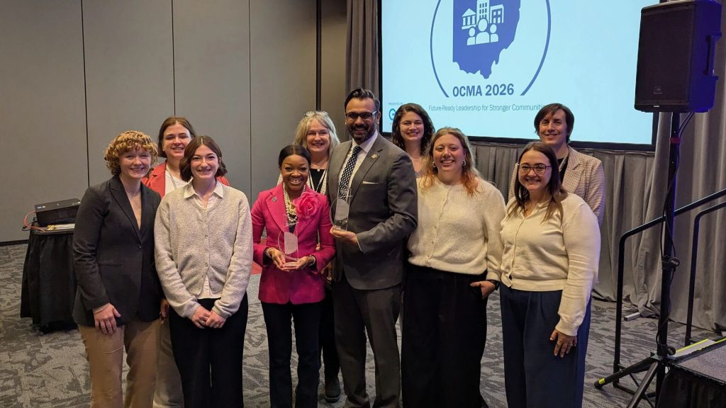 Group of nine adults posing at a conference event, with two people holding glass awards in front of a screen displaying “OCMA 2026 Future-Ready Leadership for Stronger Communities.”
