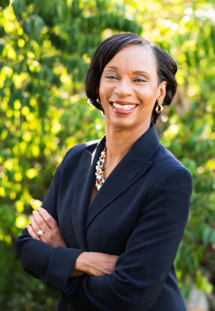 Professional headshot of a woman smiling with her arms crossed, standing outdoors in front of greenery.